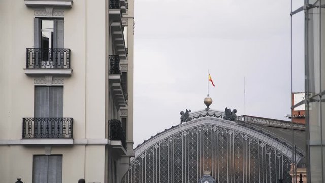 View of Madrid train station. Spanish flag in the wind. Large clock is seen in the background. People are passing by. Locked down real time medium shot