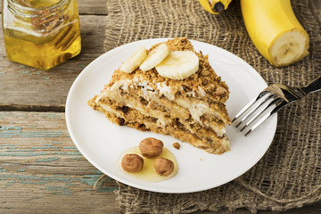 Fresh homemade banana puff cake with biscuit, sour cream, banana slices, honey on a plain gray wooden background in horizontal design. A bunch of bananas in the background.