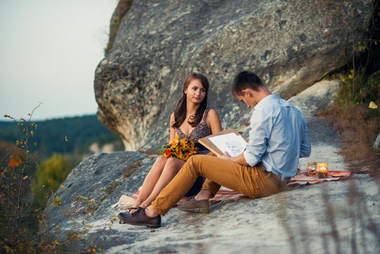 Young lovely couple on a romantic open-air date. A young man paints a portrait of his beloved girl sitting on a rock.