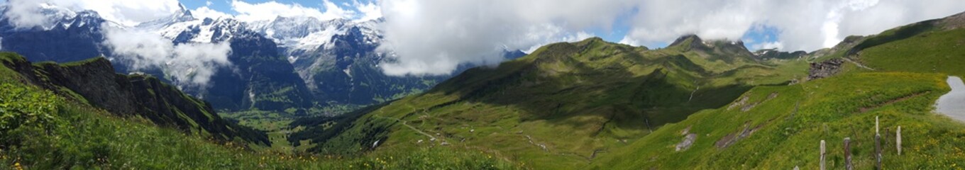 Beautiful panorama of a hidden valley surrounded by green hills, snow-covered mountains, and low clouds. View from First in Jungfrau region in Switzerland.