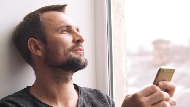 Young man looking into his iphone with positive thoughts, watching outside through the window 