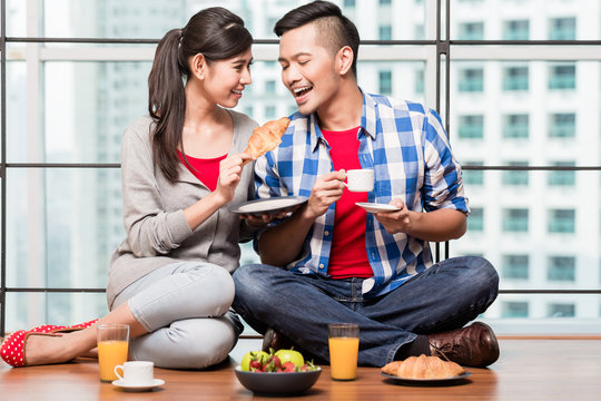 Young indonesian couple, woman and man, having breakfast in their apartment in front of city skyline