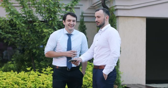 Two cheerful business men laughing smiling talking holding glass phone showing jike outdoor standing break boys guys classic clothing shirt gesturing meeting formal friends relax show elegance drink