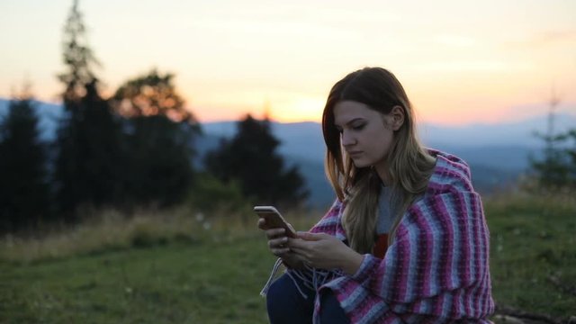 Attractive girl woman sitting using holding mobile smart phone wrapped blanket evening sunset setting sun colorful sky peaks mountains background social networking online tapping side view portrait