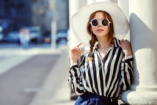 Outdoor portrait of young beautiful girl posing in street. Model wearing stylish sunglasses, hat, stripped black-white blouse. City lifestyle, female fashion concept. Copy, empty space for text
