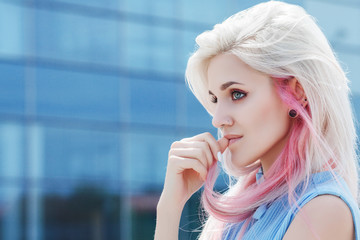 Outdoor close up portrait of young beautiful girl with makeup, long blond, pink hair. Model looking aside. Blue glass building on background. Sunny day light. Copy, empty space for text - obrazy, fototapety, plakaty