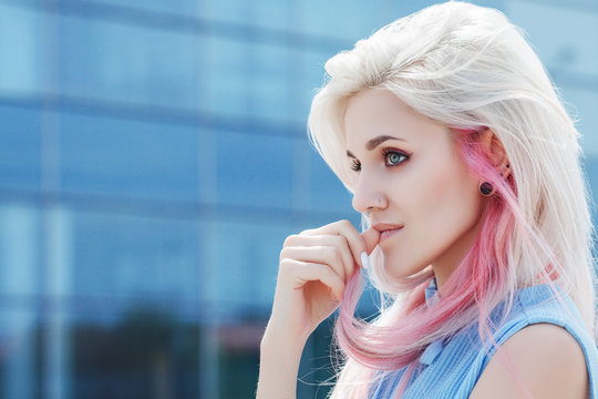 Outdoor close up portrait of young beautiful girl with makeup, long blond, pink hair. Model looking aside. Blue glass building on background. Sunny day light. Copy, empty space for text