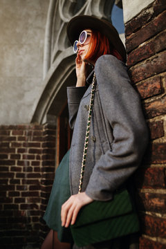 Outdoor close up portrait of young beautiful fashionable woman posing in street. Model wearing stylish hat, white round sunglasses, gray coat, with green quilted bag. Female fashion concept. 
