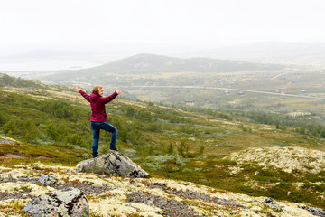 Obraz premium Young adult person looking out over foggy mountain view with small village and road. Arma out and a happy face. Location Vasstulan, Norway.