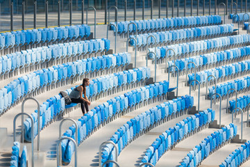 Naklejka premium Sexy girl posing at stadium with the backpack. Fitness girl with a sports figure in black leggings sitting on the seat in the stadium. Sport woman posing at stadium. Girl after sport day