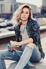 Fototapeta premium Fashionable beautiful young girl sits on the curb on a warm sunny spring evening in a city street with a skateboard and glasses. Vertical close-up portrait