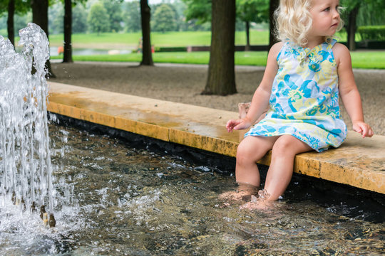 Cute little girl dipping her feet in a fountain at the park on a summer day