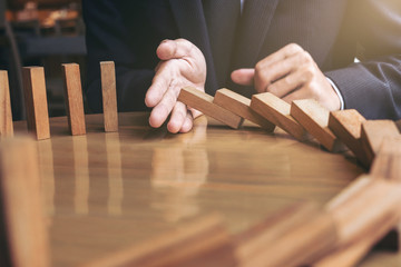 Close up of businessman hand Stopping Falling wooden Dominoes effect from continuous toppled or...