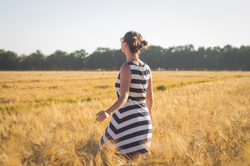 young woman in striped dress walking and having good time in summer wheat fields