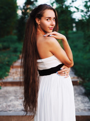 Young brunette model at the white dress staying at the stairs with green trees and looking back