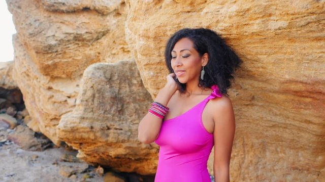Portrait of black people, pretty happy young african american woman looking at camera and smiling. Rocks beach. Sexy girl in pink swimsuit and jewelry enjoying nature. Slow motion.