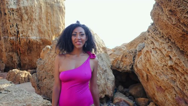Portrait of black people, pretty happy young african american woman looking at camera and smiling.Rocks beach.Sexy mixed race girl in pink swimsuit and jewelry enjoying nature.Slow motion.