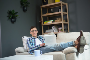 Happy young man lying on sofa, working on laptop. home office, freelancer in casual clothes working on project.