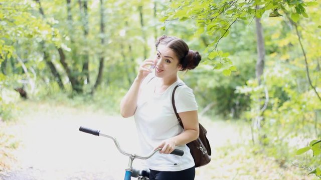 An amusing girl with a backpack near the bicycle is talking on the phone