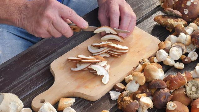 Nice wild mushrooms are cut with knife into small pieces on wooden chopping board. Man hands cut boletus slice on wooden table