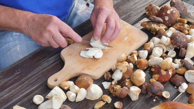 Nice wild mushrooms are cut with knife into small pieces on wooden chopping board. Man hands cut boletus slice on wooden table