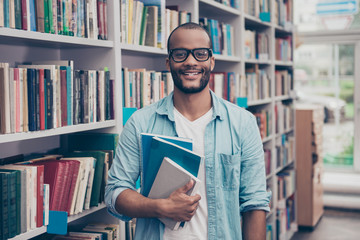Young cheerful attractive successful african nerdy student is standing with books in the school...