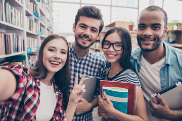 Selfie time! Four international students with beaming smiles are posing for selfie shot, caucasian attractive lady is taking, in school library building. Gathered, cheerful, smart and successful youth - obrazy, fototapety, plakaty