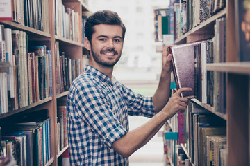 Cheerful clever young attractive brunet bearded student bookworm is studying in the ancient university library, putting vocabulary on the book shelf, smiling, wearing casual checkered shirt © deagreez