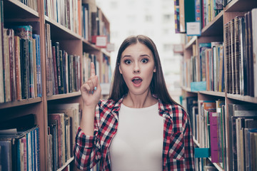 Eureka! Have an idea! Success concept. Young cute female student is standing in the ancient university`s library between book shelves, wearing casual outfit, pointing with finger up, shocked