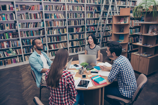 International group of four focused clever young students bookworms studying in the ancient university library, sit at the table with books and devices, talk, discuss the project, in casual outfits