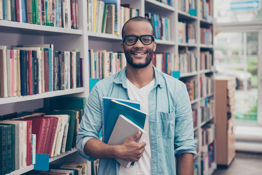 Young cheerful attractive successful african nerdy student is standing with books in the school library archive room, many tomes of ancient textbooks on shelves on the background behind