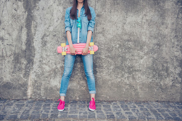 Cropped shot of cool young hipster teen in jeans outfit and pink shoes, leaning the concrete wall, holding skate board, copy space near