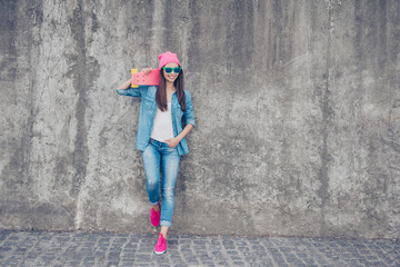 Cool swag culture. Young attractive hipster girl stands with pink skate board on concrete wall`s background, in denim jeans outfit, pink hat and shoes, holds a long board