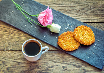 Breakfast or snack – cup of freshly brewed coffee, sesame cookies, pink lisianthus  flower and black stone tray on wooden surface