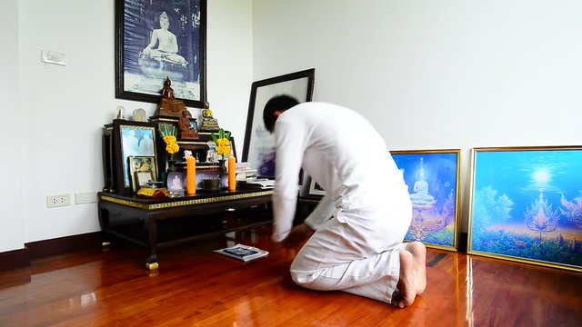 Man kneeling and making prostration to the triple gem at a set of altar table.Buddhist in white dress practicing self discipline by observing the five precepts on buddhist holy day,religion concept.