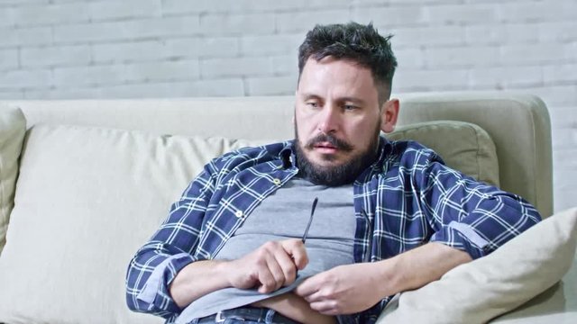 Zoom in of depressed man with beard and messy hair sitting on sofa and fidgeting, then drinking water from glass given by unrecognizable psychotherapist