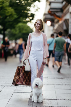 Beautiful and fashionable young woman with shopping bags and white dwarf poodle standing on street, smiling and looking at camera