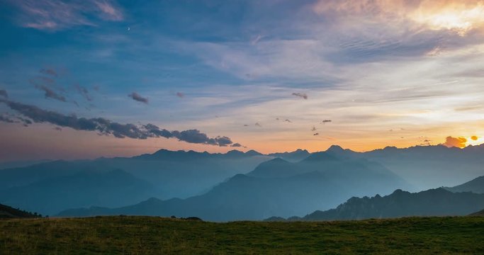 Day to night time lapse from high up on the Alps. Colorful sunset over mountain peaks and fog in the valleys below, moving clouds, setting moon and rotating stars.