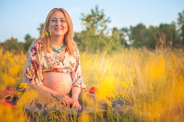 9 months pregnant woman with mehandi pattern ornament belly sitting in yellow grass and smiling on sunset lights. Waiting for baby. Pregnancy concept