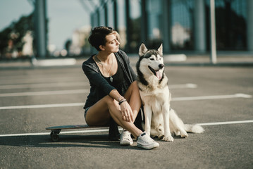 Naklejka na ściany i meble Young beautiful woman on the street hugging her husky dog sitting on a skateboard or longboard 