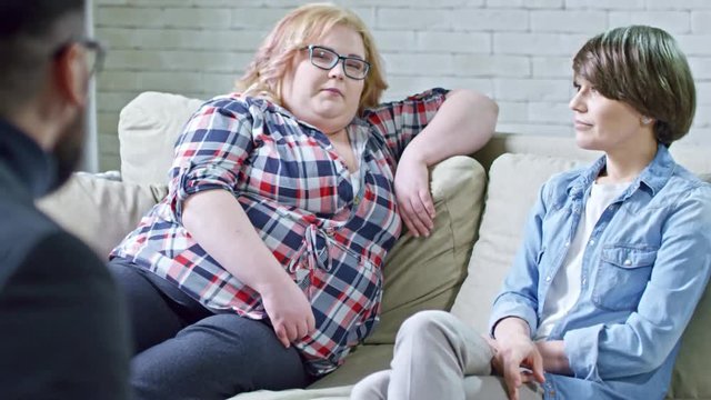 Zoom out of annoyed young woman and her overweight friend sitting on sofa and listening to male psychotherapist talking and gesturing
