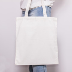 Dziewczyna w niebieskich dżinsach trzyma pustą bawełnianą torbę ekologiczną, makieta projektu. Ręcznie robiona torba na zakupy dla dziewcząt - 169684216