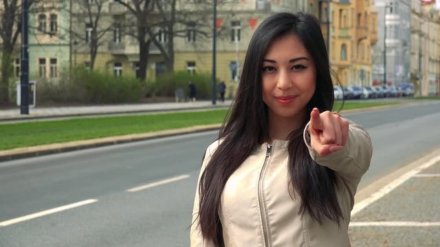 A young Asian woman points a finger at the camera and nods in a street in an urban area