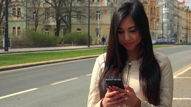 A young Asian woman works on a smartphone with a smile in a street in an urban area