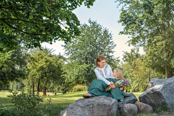 Fototapeta premium Family mom with daughter in vintage retro style linen dresses sitting on a stone rock in the park forest with bouquet of flowers looking eyes to eyes and smiling