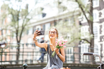 Obraz premium Młoda kobieta co selfie zdjęcie z bukietem różowych tulipanów siedzi na płocie w pobliżu kanału wodnego w mieście Amsterdam