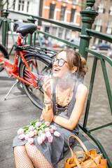 Naklejka premium Młoda uśmiechnięta kobieta pali papierosa siedząc z kwiatami i rowerem na moście w mieście Amsterdam