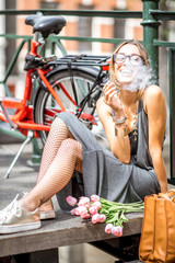 Naklejka premium Młoda uśmiechnięta kobieta pali papierosa siedząc z kwiatami i rowerem na moście w mieście Amsterdam