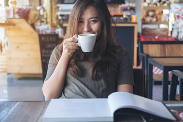 Closeup image of a beautiful Asian woman reading a book while holding and drinking hot coffee in modern cafe
