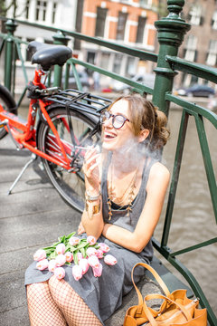 Young smiling woman smoking a cigarette sitting with flowers and bicycle on the bridge in Amsterdam city